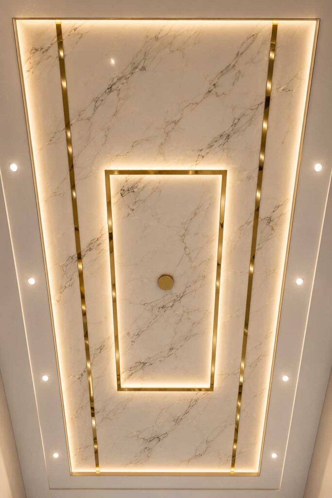 Luxury marble false ceiling design featuring gold metal inlay strips, warm LED cove lighting, recessed spotlights, geometric rectangular patterns, and a modern premium interior style.