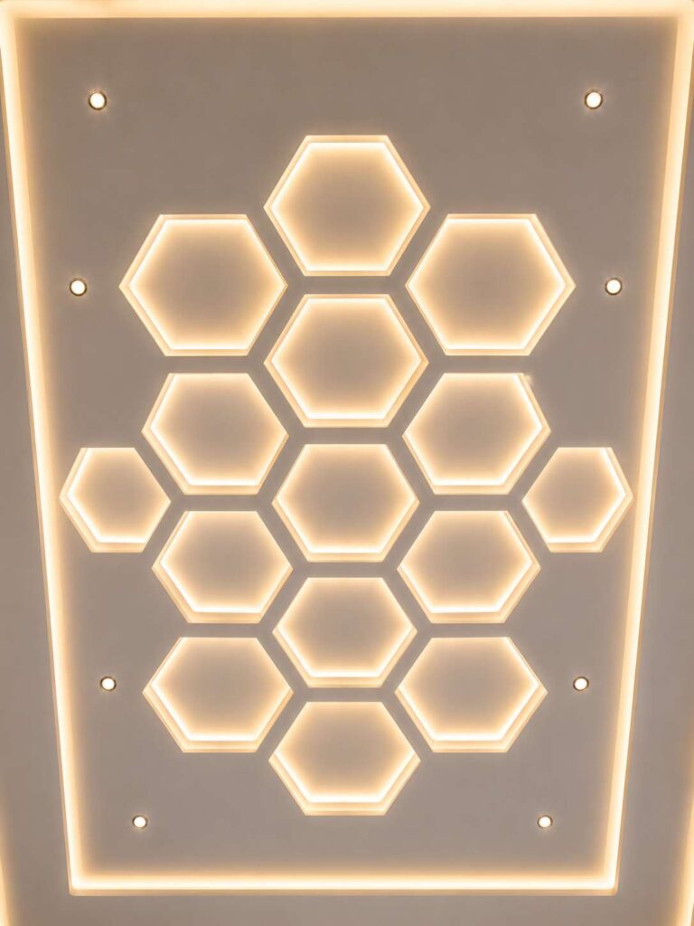 Modern hexagon false ceiling design with honeycomb geometric patterns, warm LED backlighting, recessed spotlights, cove lighting, and contemporary minimalist interior styling.